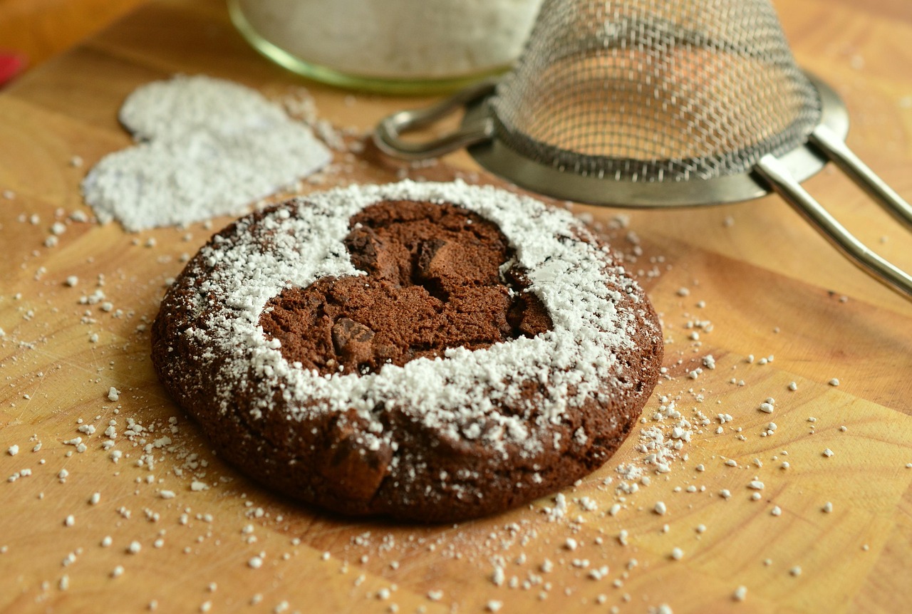 The image shows a rich chocolate cookie dusted with powdered sugar. A heart-shaped stencil in the centre reveals the cookie’s surface beneath the sugar, surrounded by scattered icing sugar and a metal sifter on a wooden board — evoking a warm, homemade fall baking vibe.